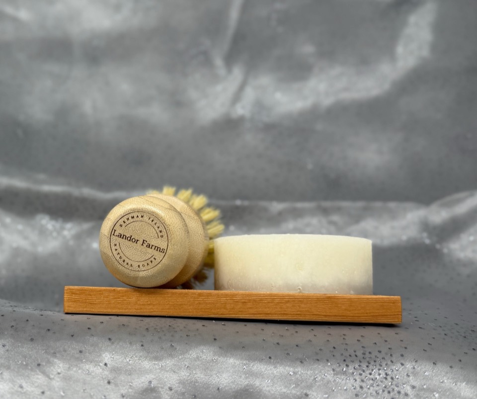 Landor Farms dishwashing brush lying beside a round cream soap bar on a wooden soap stand, on grey satin