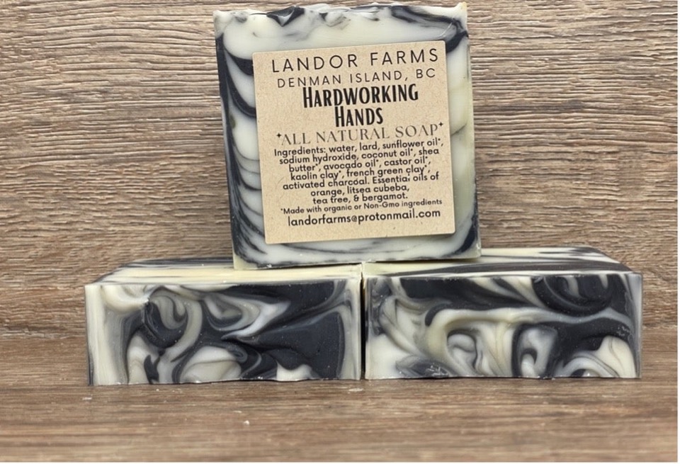 Three Hard Working Hands soap bars with black and cream swirls on a wood backdrop; the top bar has a Landor Farms label