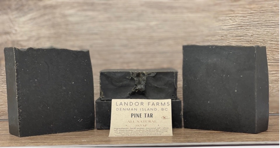 Four black Pine Tar soap bars on a wood backdrop, with a Landor Farms Pine Tar label in front