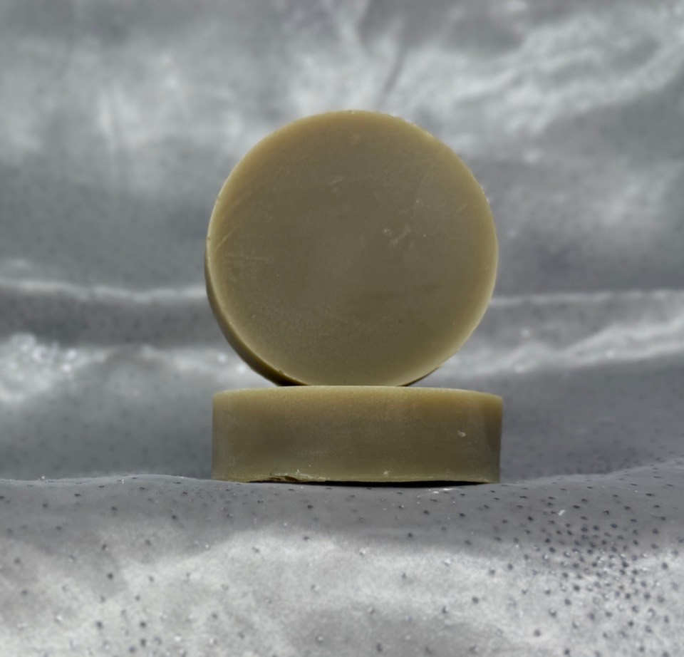 Shampoo + Conditioner Set: two round olive-green shampoo bars, one on edge and one lying flat in front of it, on grey satin